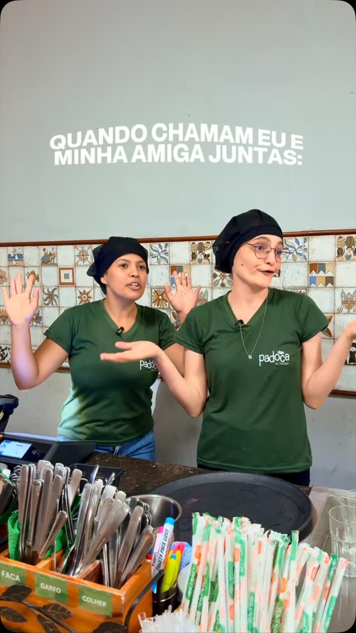 Duas funcionárias da Padoca de camisa verde acenando atrás do balcão, com a frase quando chamam eu e minha amiga juntas escrita à mão pela Padoca.