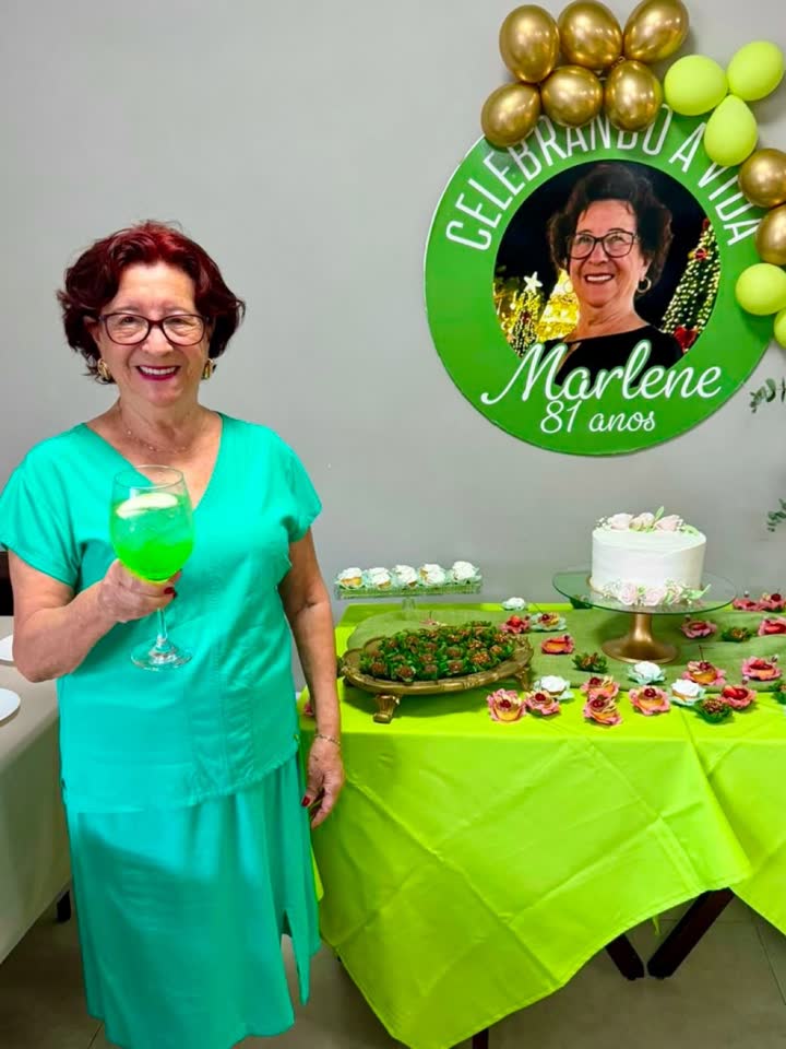 Dona Marlene, de verde, segurando um drink ao lado do painel redondo escrito Celebrando a vida, Marlene, 81 anos.