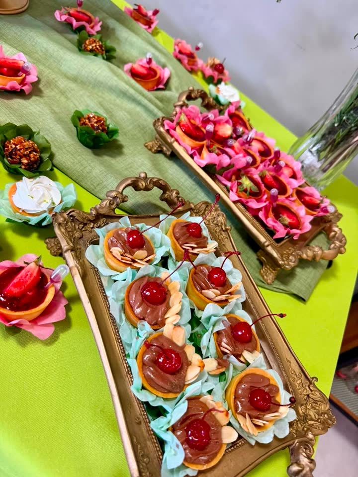 Mesa da festa de 81 anos montada na Padoca: bandejas douradas com tortinhas de cereja e de morango sobre toalha verde-limão.