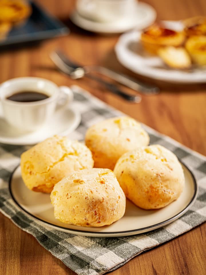 Prato de pães de queijo dourados sobre guardanapo xadrez, com uma xícara de café preto ao lado.