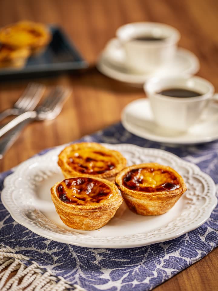 Três pastéis de Belém em um prato de porcelana, com café e talheres sobre toalha estampada.