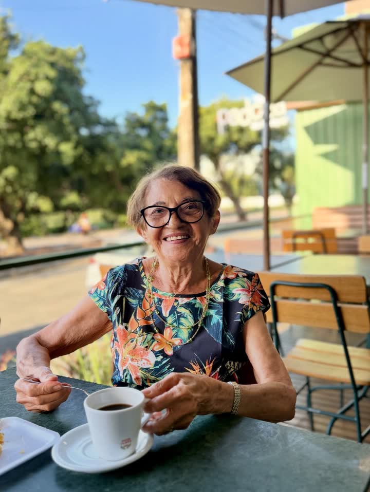 Senhora sentada sozinha na mesa da calçada da Padoca, embaixo do guarda-sol, tomando um café.